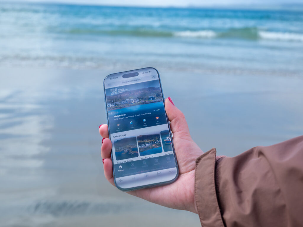 Discover Arisaig app on iPhone in a person's hand in front of the shoreline